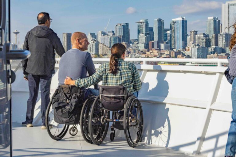 Two people in wheelchairs and another standing person look out from a boat deck toward a city skyline under a blue sky. The sun casts shadows, and the city buildings rise in the background.