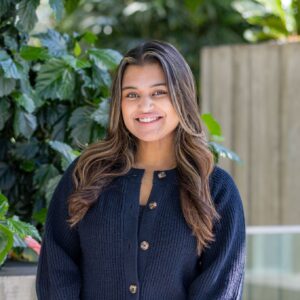 A young woman with long brown hair wearing a navy blue sweater with wooden buttons stands outdoors, smiling in front of green leafy plants and a wooden fence.
