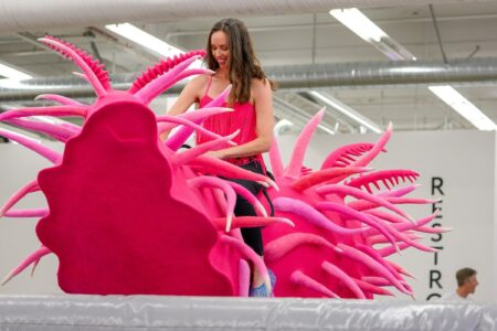 A woman in a pink top interacts with a large, pink, spiky, abstract sculpture in an indoor space with bright lighting and visible ductwork. Other people are present in the background.