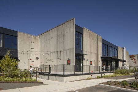 A modern building with a concrete exterior, large windows, and a metal ramp entrance under a clear blue sky. There are small trees and landscaped plants in front, and a few people walking near the entrance.