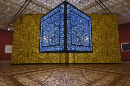 A large, blue, intricately patterned light box hangs in a gallery, casting elaborate geometric shadows on the yellow walls and wood floor, creating a mesmerizing and immersive visual effect.