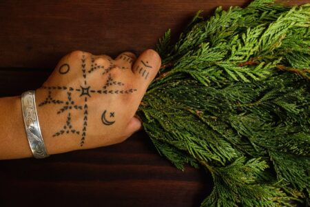 A hand with geometric and symbolic tattoos, wearing a silver bracelet, holds a bundle of green foliage on a dark wooden surface.
