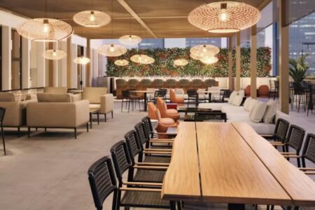 Modern rooftop lounge with wooden tables, woven chairs, cushioned sofas, hanging pendant lights, and green wall decor. Large windows offer city views. The space appears stylish, open, and inviting.