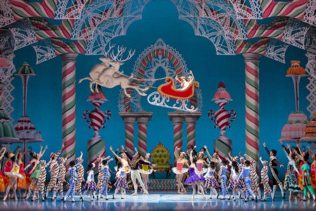 A colorful stage scene from The Nutcracker ballet features dancers in festive costumes, candy-themed decorations, and a sleigh with Santa and reindeer flying above the performers who raise their arms in celebration.
