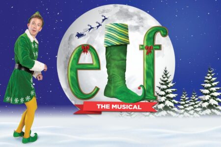 A person in a green elf costume stands in snow beside large festive letters spelling 