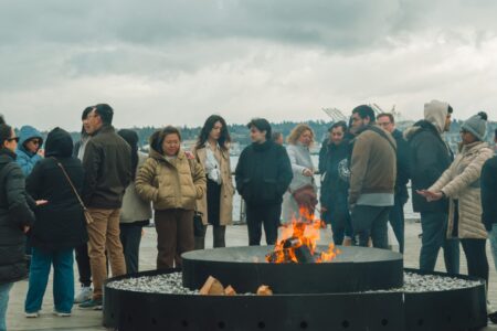 A group of people dressed in winter clothing gather around a large outdoor fire pit on a cloudy day. Smoke rises from the flames, and the waterfront and trees are visible in the background.