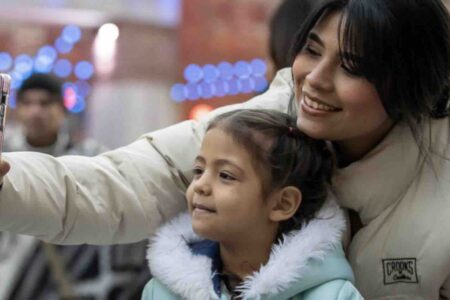 A woman and a young girl in winter jackets smile as they take a selfie together outdoors, with colorful lights blurred in the background.