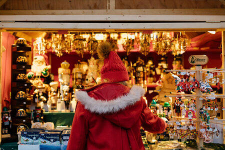 A person in a red coat and pom-pom hat browses festive ornaments and nutcrackers at a warmly lit holiday market stall decorated with hanging golden ornaments.