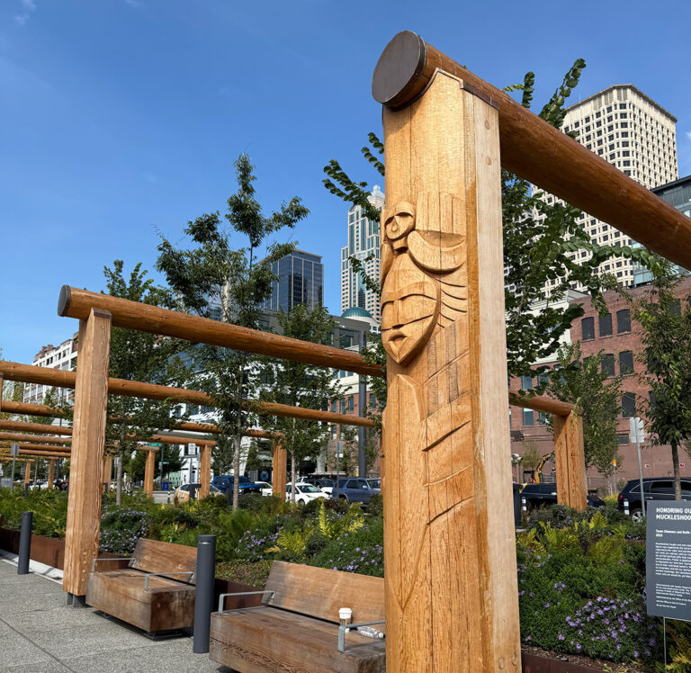 A wooden pergola with Indigenous-style carvings stands in an urban park area with benches below, greenery, and tall city buildings in the background under a clear blue sky.