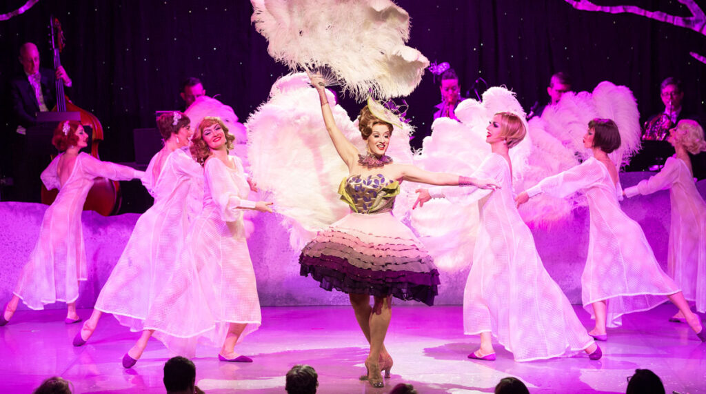 A group of performers in glamorous costumes dance on stage at one of the city’s top music venues. The central figure wears a ruffled dress and holds large feather fans, while backup dancers in pink dresses pose behind her under the bright lights.
