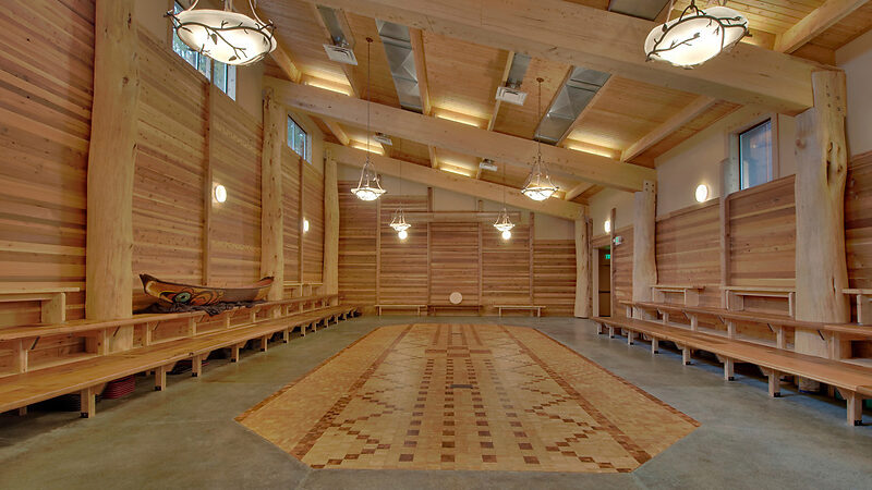 A spacious wooden room with high ceilings, decorative hanging lights, long benches along the walls, and a patterned wooden floor. A carved canoe is positioned on the left side near the wall.
