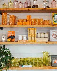Shelves display orange and yellow glassware, books, and candles. The top shelf has jars and bottles, the middle shelf has pitchers, books, and candles, and the bottom shelf holds green glass jars and pitchers, with a plant on the left.