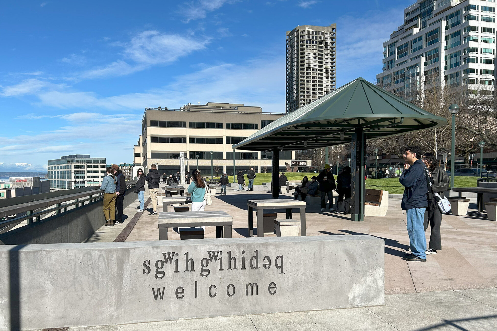 A sunny outdoor plaza with people standing and sitting near tables and a pavilion. A concrete wall in the foreground says "welcome" with upside-down and mirrored text above it. Modern buildings and blue sky are in the background.