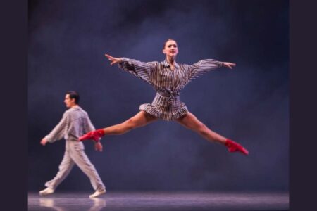 A dancer in a striped outfit and red socks leaps with arms and legs extended, while another dancer in similar striped clothing walks in the background on a smoky, dimly lit stage.