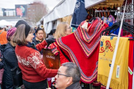 People browse colorful embroidered clothing at an outdoor market stall. A woman in a red sweater holds a bright red shawl. Other shoppers look at items, and festive decorations are visible in the busy market setting.