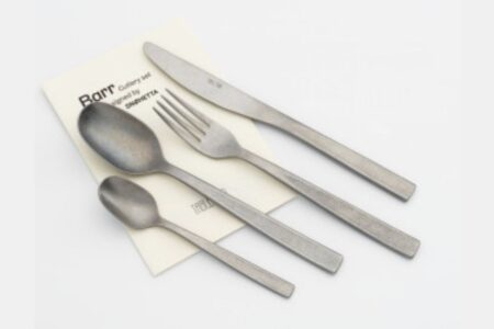 A set of four stainless steel utensils—a knife, fork, tablespoon, and teaspoon—lies on a folded cream-colored paper with text, arranged neatly on a white surface.