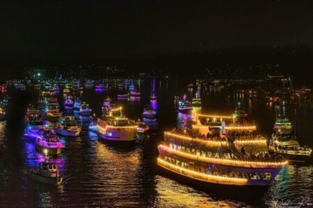 A fleet of boats and yachts decorated with colorful, glowing holiday lights float on a dark body of water at night, creating a festive and vibrant scene.