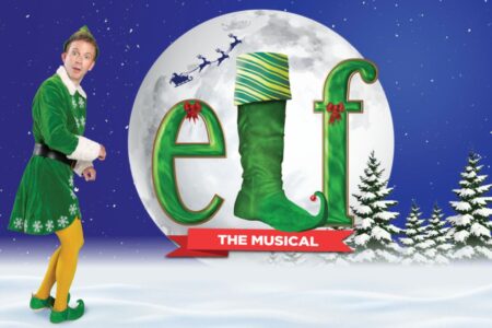 A man dressed as an elf stands in snow beside the large, festive word 