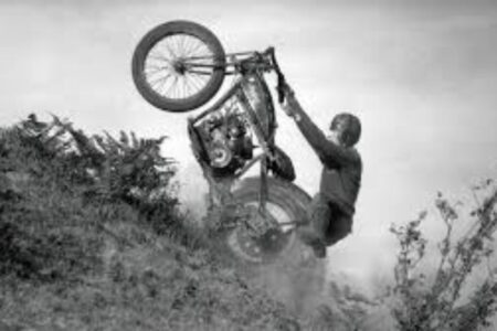 A person riding a motorcycle up a steep hill loses balance, causing the front wheel to lift off the ground. Dust and plants are visible around the motorcycle. The image is in black and white.