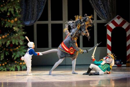 A ballet scene features a dancer in a mouse king costume brandishing a sword at a performer dressed as a nutcracker soldier on the floor, while a child bunny character tugs at the mouse king's tail.