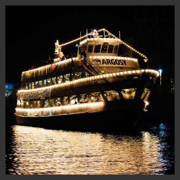 A large Argosy cruise boat decorated with string lights is illuminated at night on the water, reflecting its glow onto the surface.