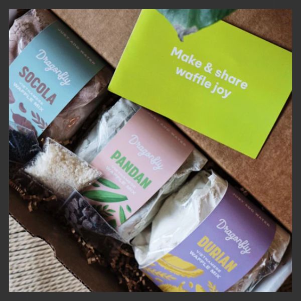 A box containing three Dragonfly waffle mix packs labeled Socola, Pandan, and Durian, next to a green card that says, "Make & share waffle joy.