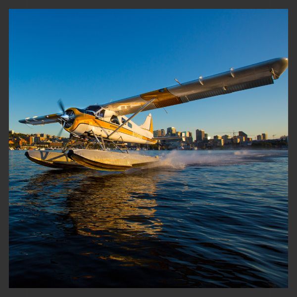 A yellow and white seaplane takes off from a calm body of water, with a city skyline and buildings visible in the background under a clear blue sky.