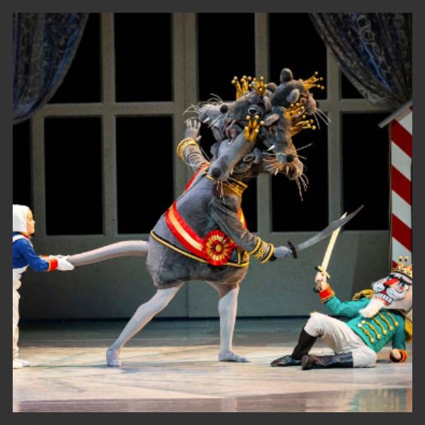 A ballet performance featuring a dancer in a large, multi-headed rat costume with a red sash and crown, holding a sword, facing a performer dressed as a toy soldier with a nutcracker mask. Another dancer stands to the side.
