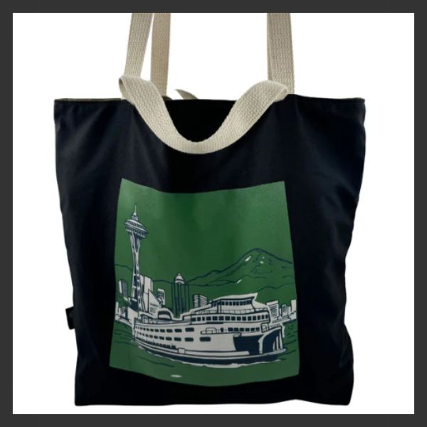A black tote bag with thick cream-colored handles features a green and white illustration of the Seattle skyline, including the Space Needle, a ferry, mountains, and water.