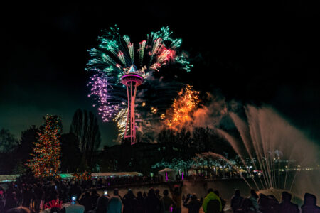 Colorful fireworks burst around the Space Needle at night, lighting up the sky above a crowd of people. A decorated Christmas tree and illuminated water fountains are also visible in the festive scene.