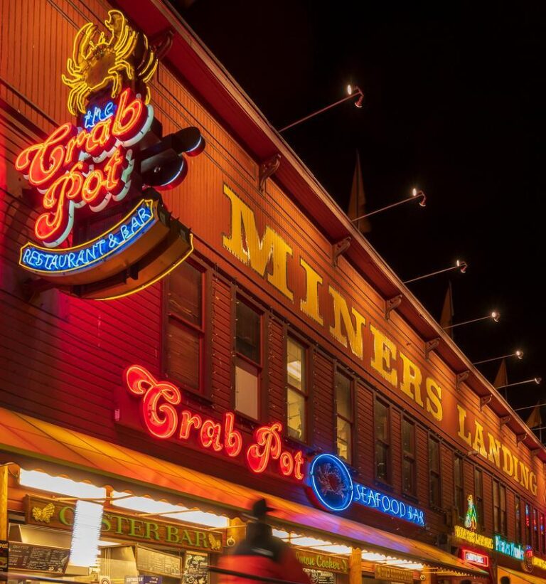 A brightly lit seafood restaurant called "Crab Pot" at "MINERS LANDING" is shown at night. Neon signs glow as people walk by, and a cyclist passes in the foreground, creating a sense of motion.