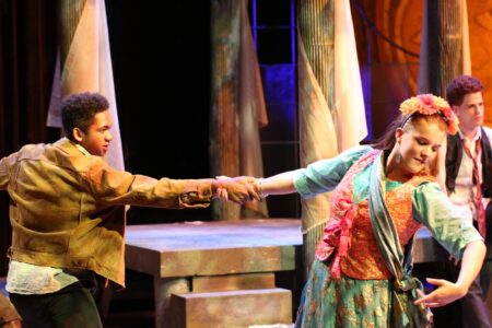 Two actors on stage hold hands mid-dance, wearing colorful period costumes. Other performers sit in the background, and the scene is lit with warm, theatrical lighting. The setting suggests a dramatic or musical play.