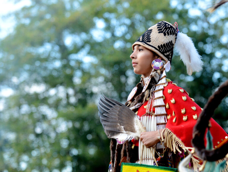A person in traditional Native American regalia, adorned with beadwork, feathers, and a decorated headdress, stands outdoors against a blurred background of trees, holding a feather fan and looking thoughtfully into the distance.