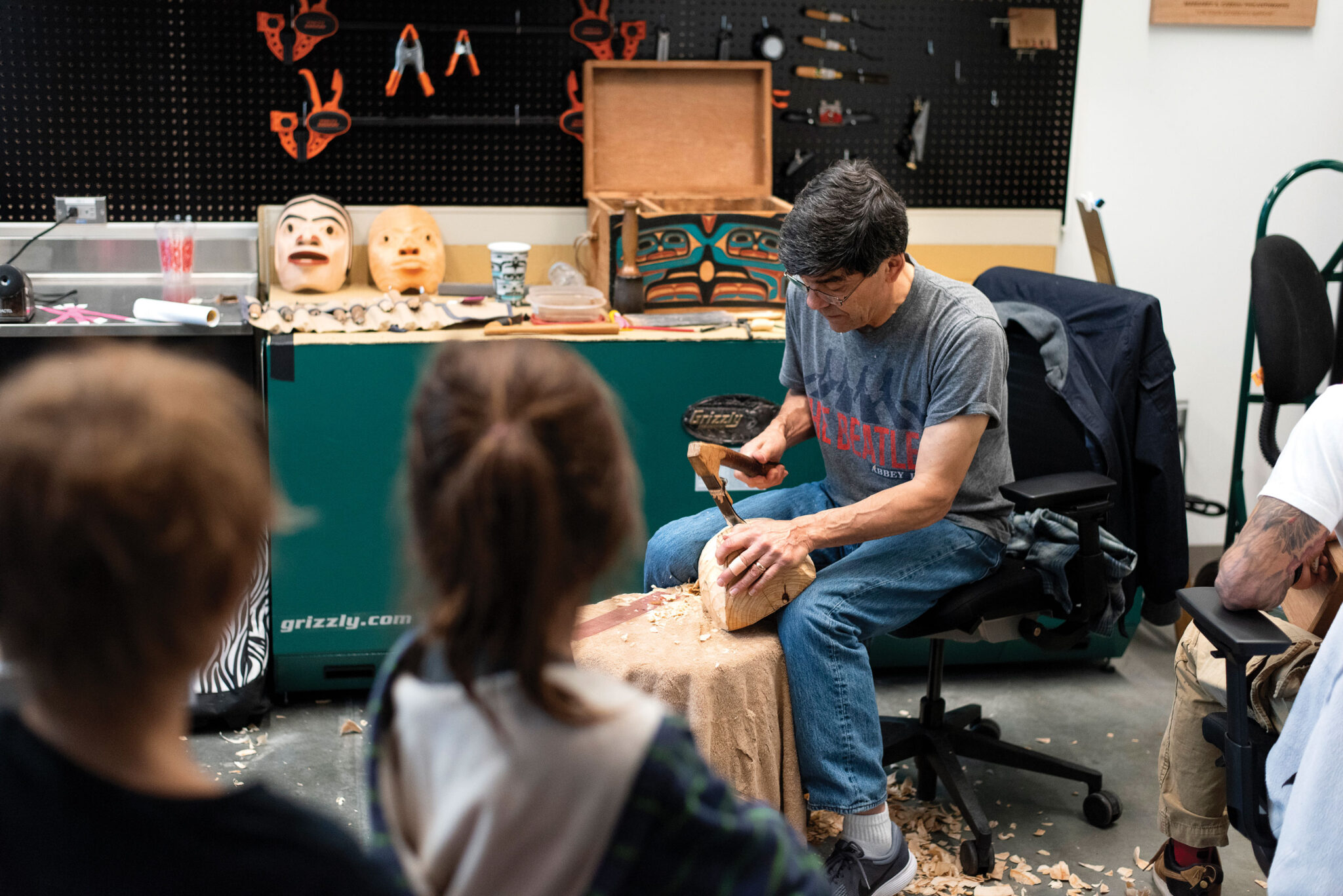 A man sits carving a wooden mask with a chisel in a workshop, surrounded by tools and completed masks, while two people watch from the foreground.