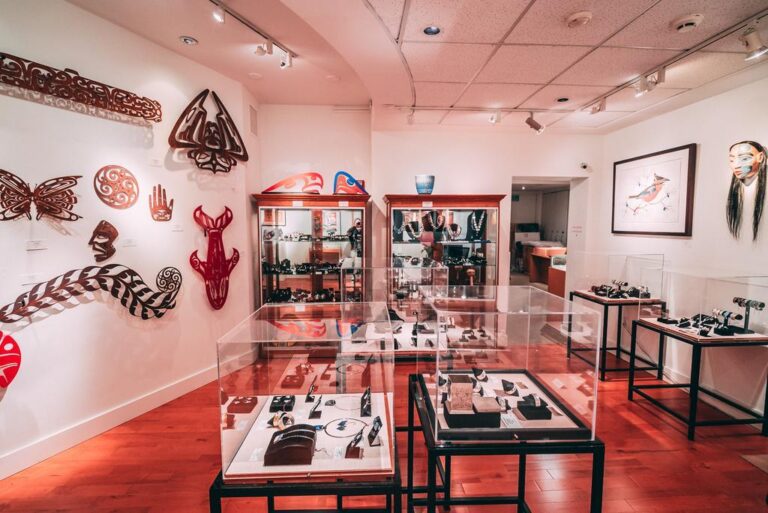 A gallery with wooden floors displays glass cases of jewelry and shelves of handcrafted items. The walls are adorned with various Indigenous art pieces, including masks, carvings, and framed artwork.