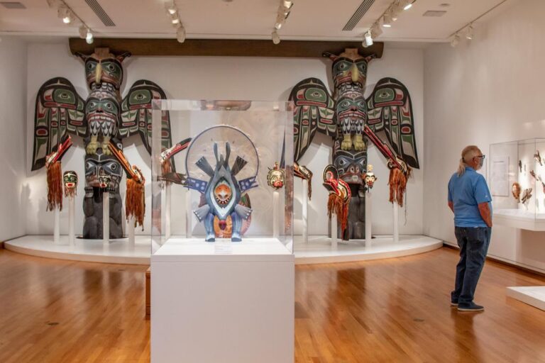 A museum gallery displays large, colorful totem poles and Indigenous masks. A man in a blue shirt and jeans stands observing the exhibits. The room has wooden floors and white walls.