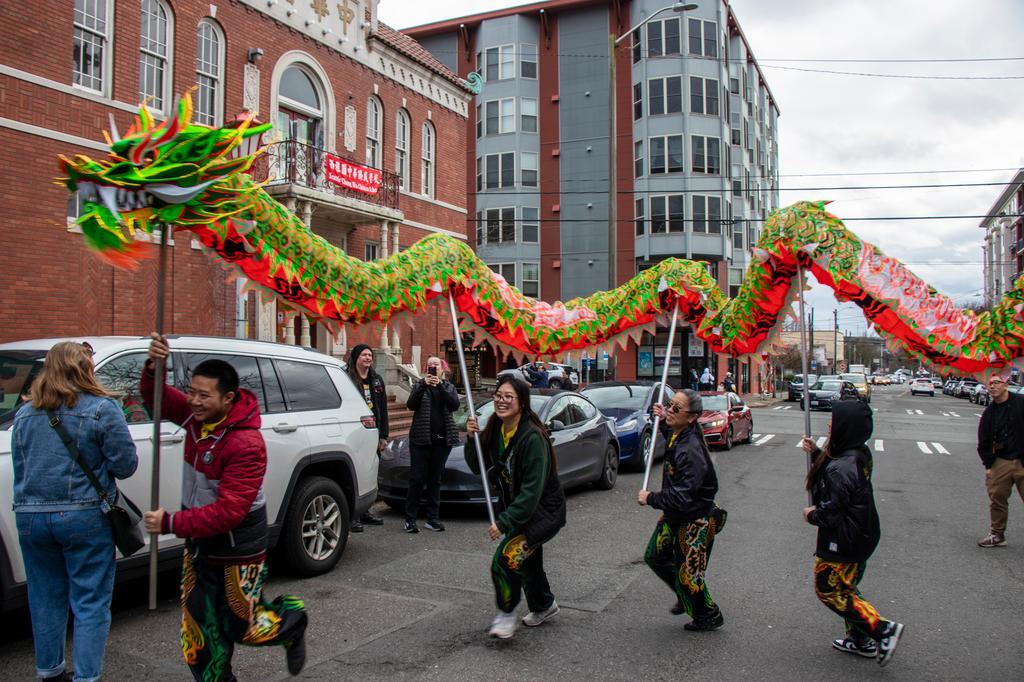 People perform a traditional dragon dance on a city street, carrying a colorful dragon costume. Bystanders watch and take photos. Brick and modern buildings line the background.