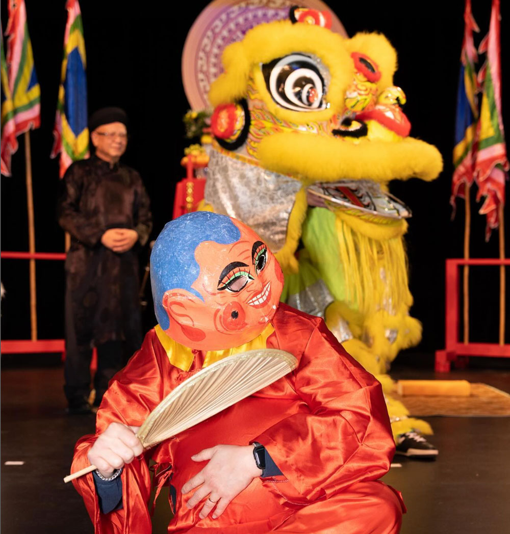 A person in a colorful Buddha mask and red robe holds a fan, sitting in front of a yellow lion dance costume with vibrant flags and another person in black traditional attire in the background.