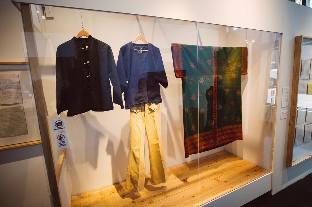 Three pieces of clothing are displayed on hangers in a glass case: a black jacket, beige pants with a blue top, and a green garment with red and gold patterns, likely traditional attire.