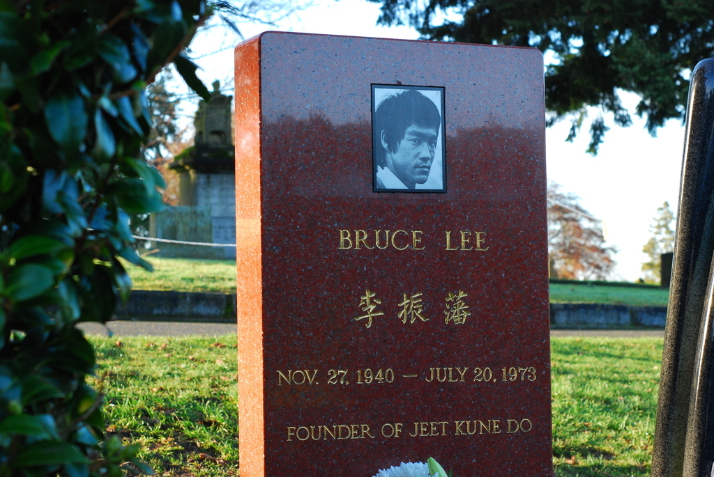 A red gravestone with a black-and-white photo of Bruce Lee, his name, Chinese characters, dates (Nov. 27, 1940 – July 20, 1973), and the words "Founder of Jeet Kune Do" engraved in gold.