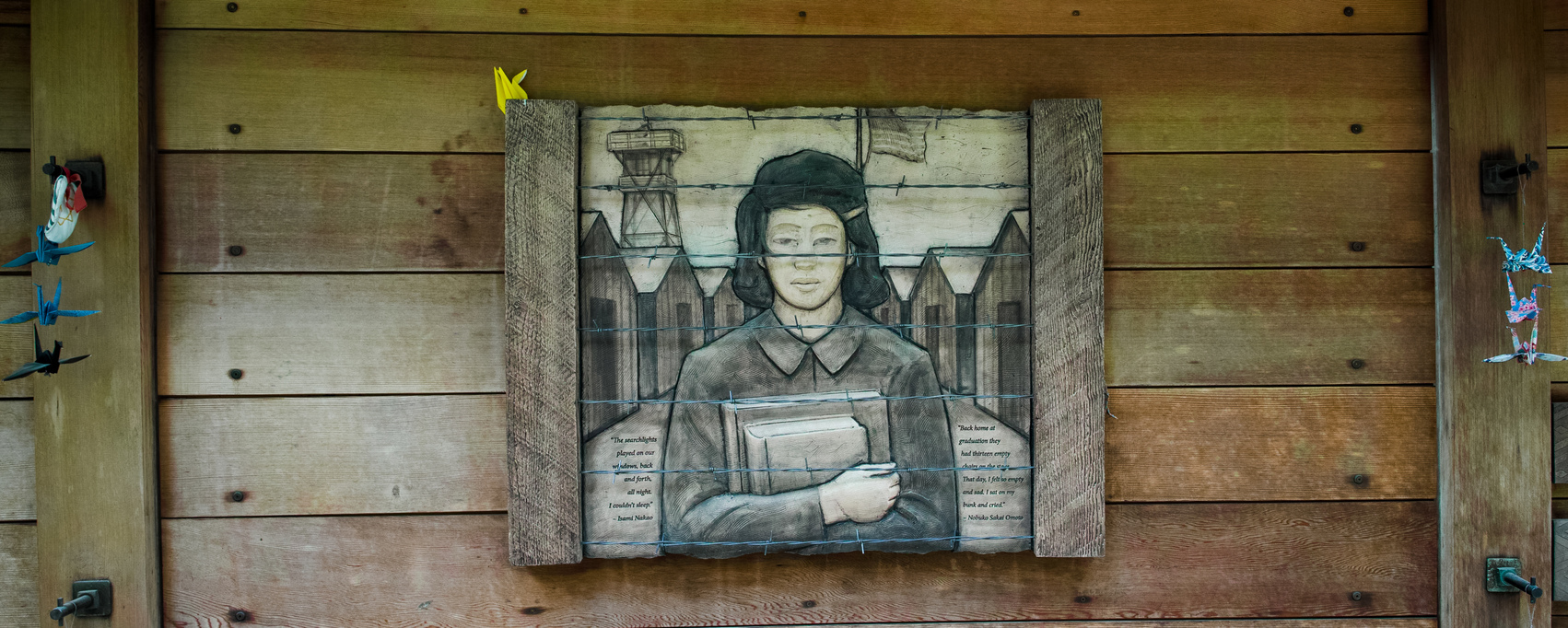 A wooden wall displays a framed illustration of a person holding books, with barbed wire and guard towers in the background, suggesting a historical or prison camp theme. Model airplanes and a Santa figure are mounted nearby.