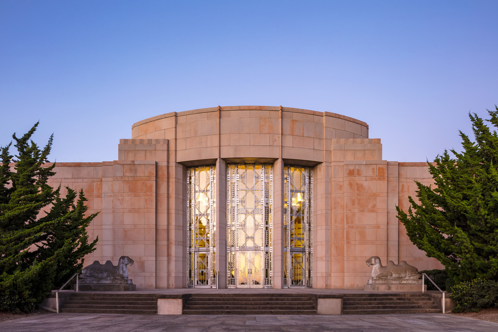 Art Deco-style building with a central rounded facade, tall decorative glass and metal doors, symmetrical stone steps, and sculpted animal figures on either side, flanked by neatly trimmed bushes at sunset.