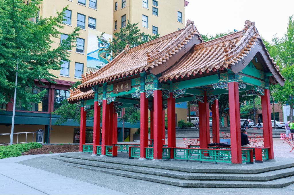 A traditional Chinese pavilion with red pillars and a tiled roof stands in a public plaza, surrounded by trees and modern buildings in the background.