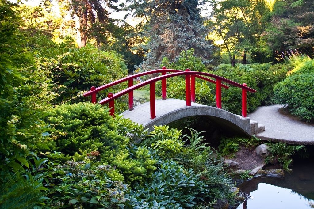 A small arched bridge with bright red railings spans over a narrow stream, surrounded by lush green foliage and trees in a peaceful garden setting.
