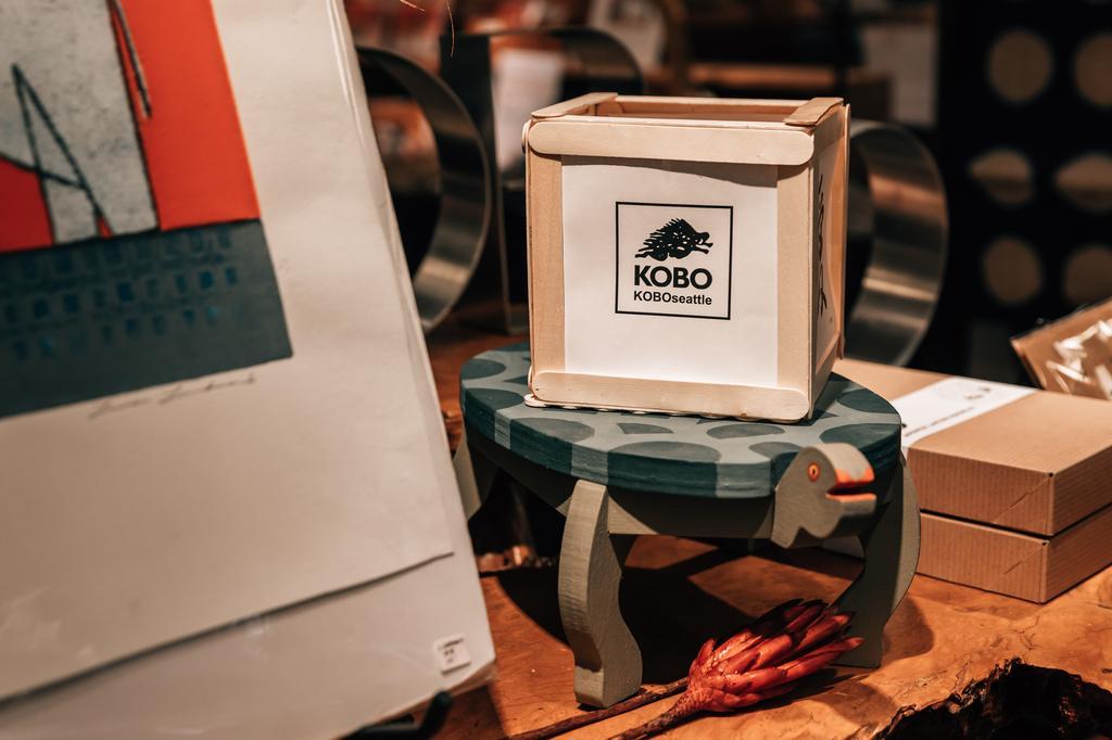 A small white box labeled "KOBO" sits on a turtle-shaped wooden stool, surrounded by art prints and wooden objects on a tabletop in a cozy, artsy shop setting.
