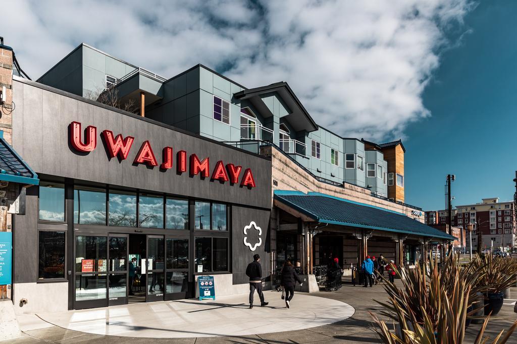 People walk near the entrance of Uwajimaya, an Asian supermarket with modern gray exterior and bold red signage, on a partly cloudy day. Contemporary apartments rise above the store.