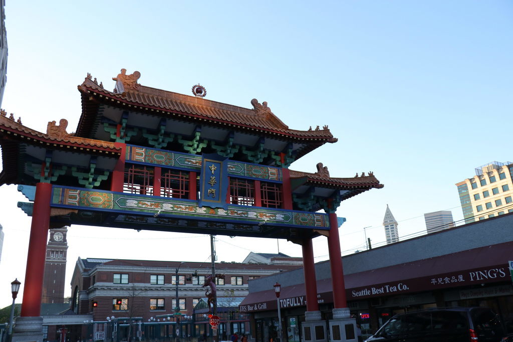 A traditional Chinese-style gate with ornate red pillars and green, gold, and blue details stands at the entrance to a Chinatown district, surrounded by city buildings and shops beneath a clear blue sky.