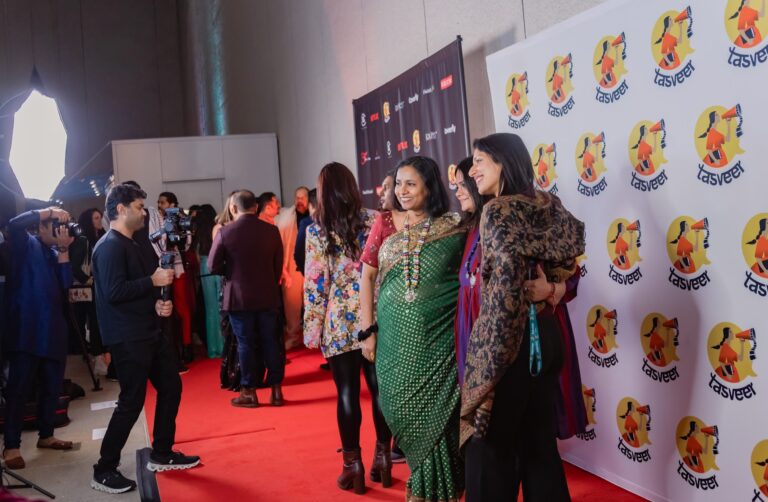 People pose for photos on a red carpet at an event, with photographers on the left and a backdrop featuring the Tasveer logo on the right. Guests wear colorful, festive attire and smile for the cameras.
