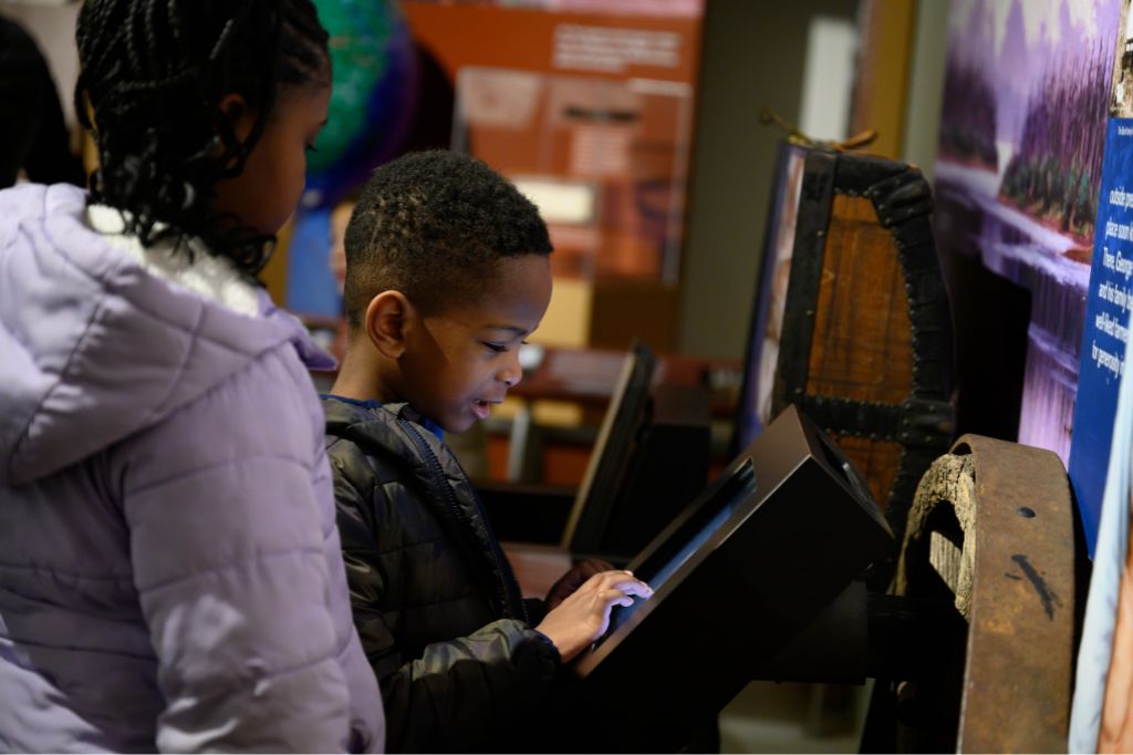 A young boy using a touchscreen display at a museum exhibit, with a girl standing beside him, both wearing jackets and looking engaged.