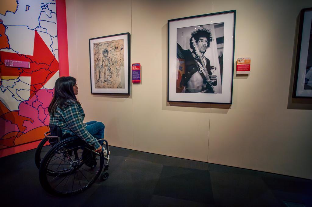 A woman in a wheelchair looks at framed art and a black-and-white photograph of a guitarist on display in a museum gallery.